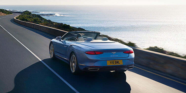 JCT600 Service Teeside New Bentley Continental GTC Azure convertible rear view in Light Windsor blue paint with 22 inch ten spoke wheels driving dynamically on an ocean road in the sunshine