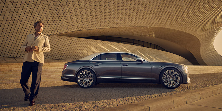 JCT600 Service Teeside Bentley Flying Spur Mulliner sedan in Rose Gold over Magnetic duotone paint parked by a curved wall with person in suit jacket standing on the left